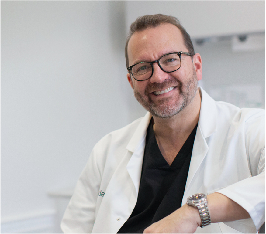 Dr Mark Hughes smiling in a bright clinical setting, wearing glasses and a white clinical coat.
